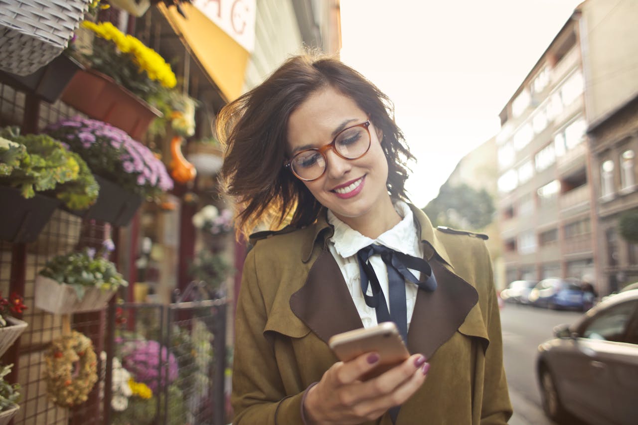 why-choose-us A smiling woman uses her phone outside a colorful urban flower shop on a sunny day.